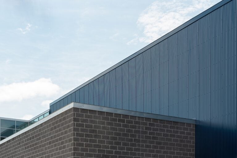 The exterior wall of a commercial building with grey brick, light gray mortar, and silver color flashing or fascia. There's a deep blue sheet metal siding wall on a high exterior wall above the brick