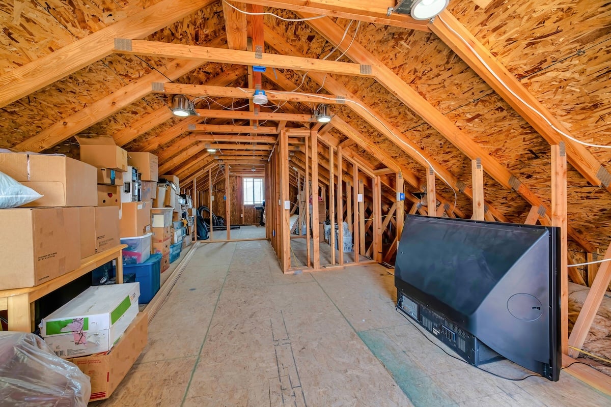Interior of the attic of house with boxes and old appliances under gable roof. Round lights are mounted on the wooden frames of the ceiling.