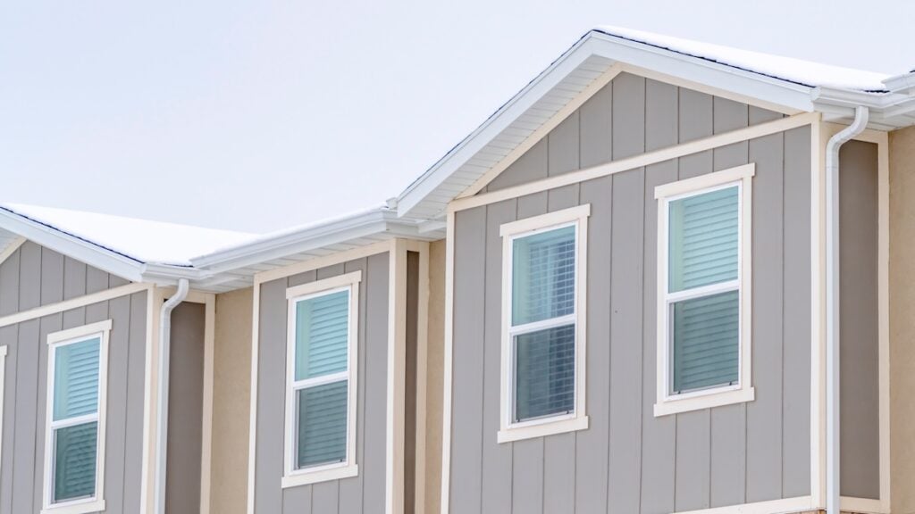 Panorama Snowy roof brick wall and vertical siding at exterior of townhome against sky. Residential landscape with view of the upper story of townhouses.